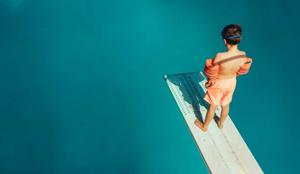 Top view of boy standing on spring board learning to dive during swimming class on a summer day. Boy learning swimming at outdoor pool.