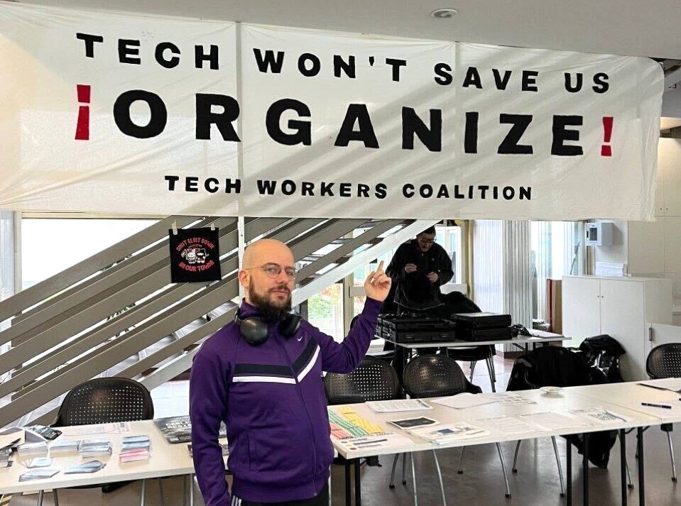 foto de Simone Robutti apontando para a placa da coalizão de trabalhadores de tecnologia que diz que a tecnologia não nos salvará - organize
